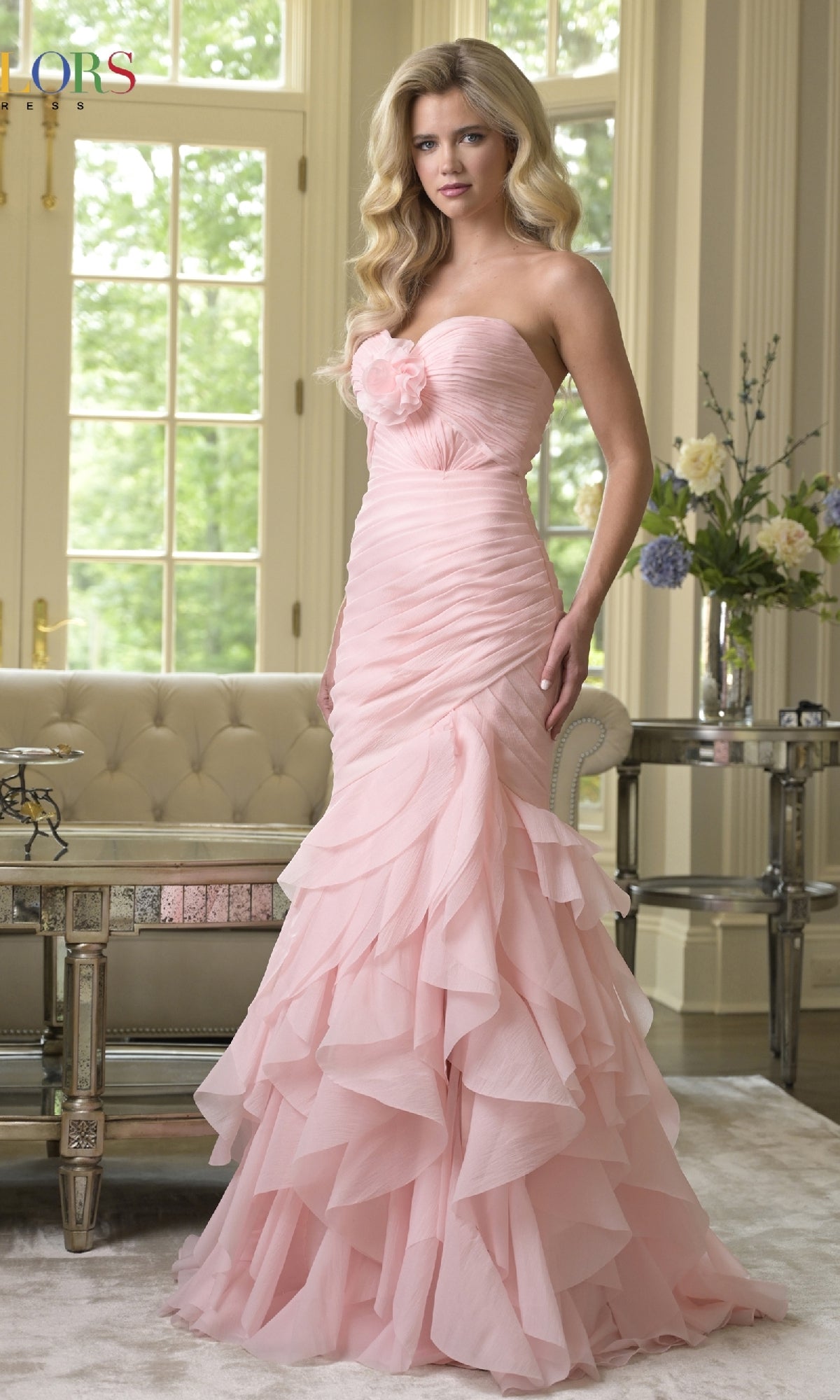 A woman stands indoors near a sofa and large windows, wearing the blush Strapless Chiffon Prom Gown: Colors Dress 3886; flowers decorate a nearby table.
